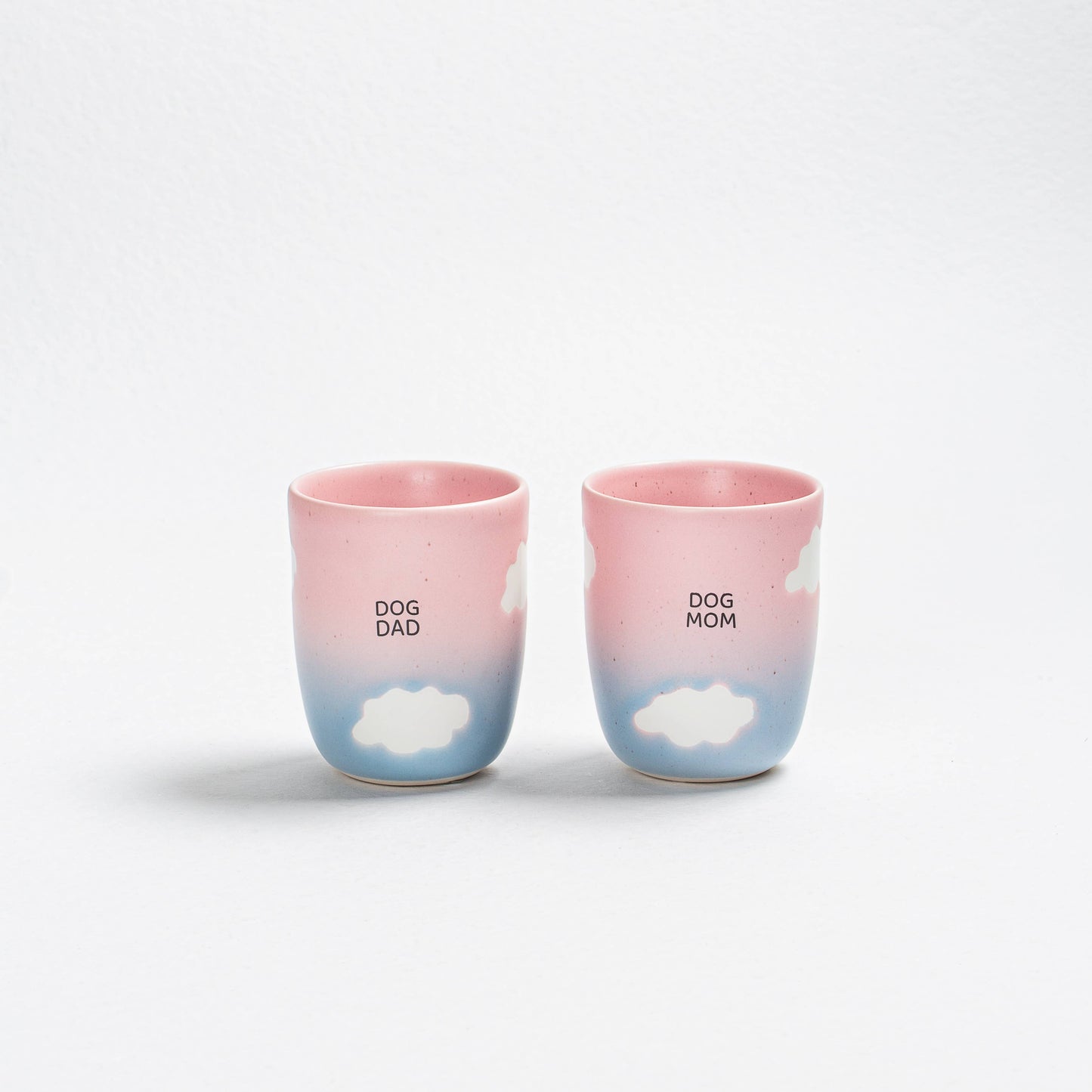 Two ceramic cups with 'DOG DAD' and 'DOG MOM' text and cloud designs on a white background.