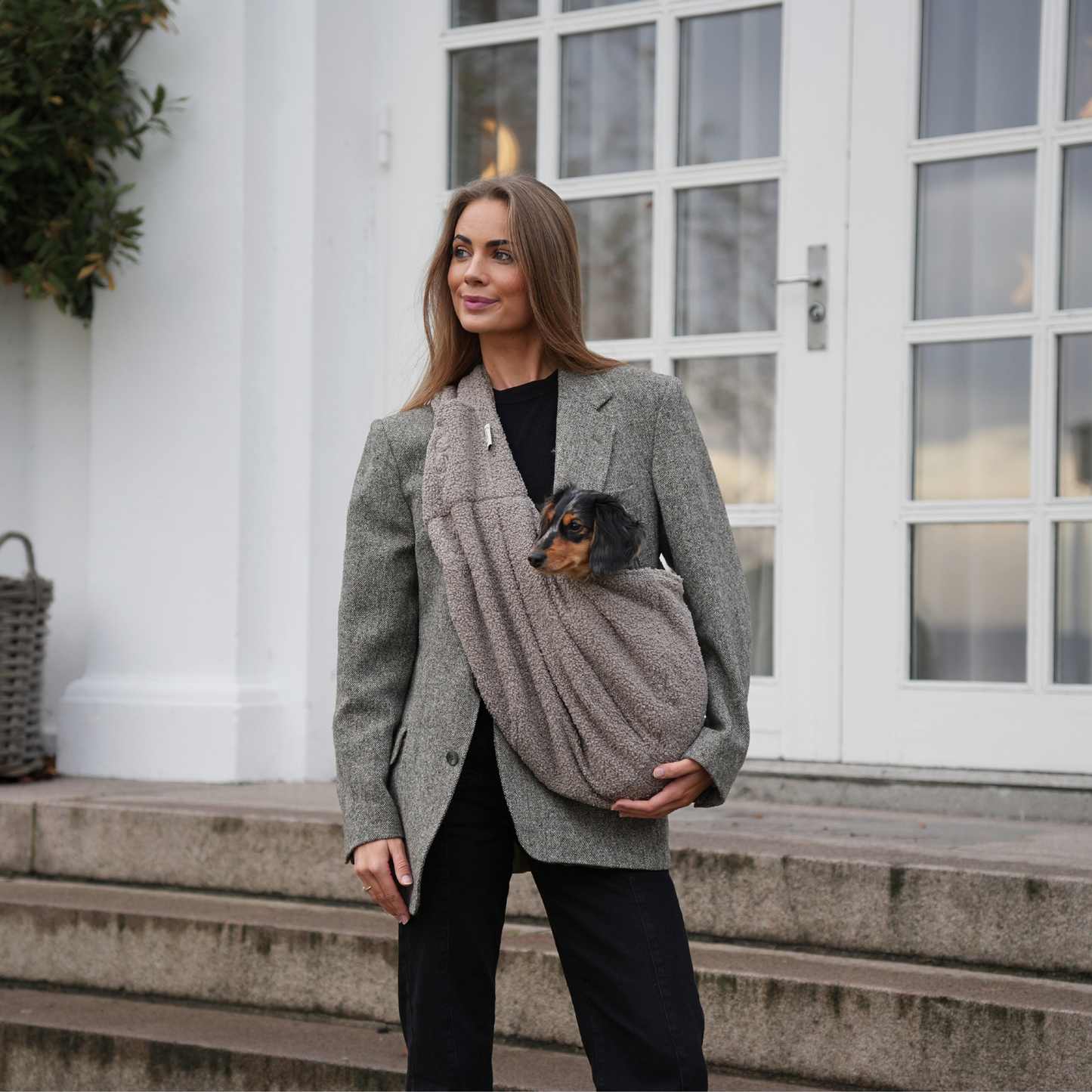 Woman holding a small dog in a sling while standing outdoors.