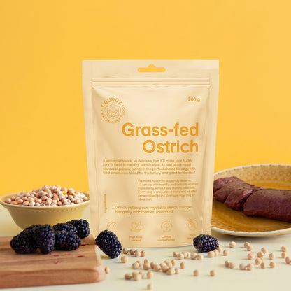 A stand-up pouch of Midol Organic Grass-fed Ostrich meat in a light beige color, styled on a white surface with a yellow background. The pouch is surrounded by ingredients including blackberries, a bowl of soybeans, and a plate of raw ostrich meat.