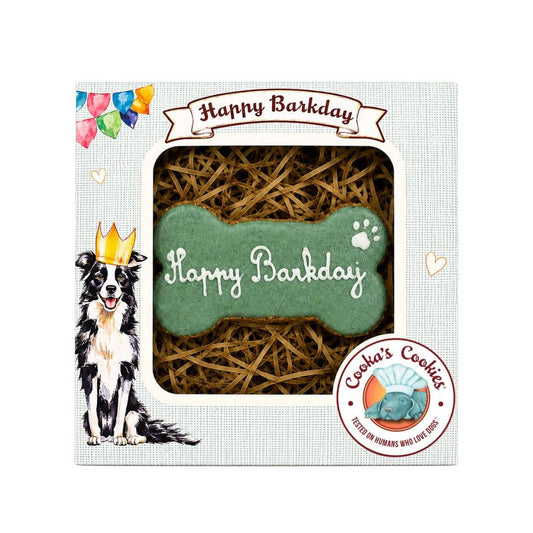 A boxed pet birthday cookie gift set for dogs on a white background. The package shows a bone-shaped green cookie with white script reading 'Happy Barkday' and a paw print, surrounded by brown shredded paper.