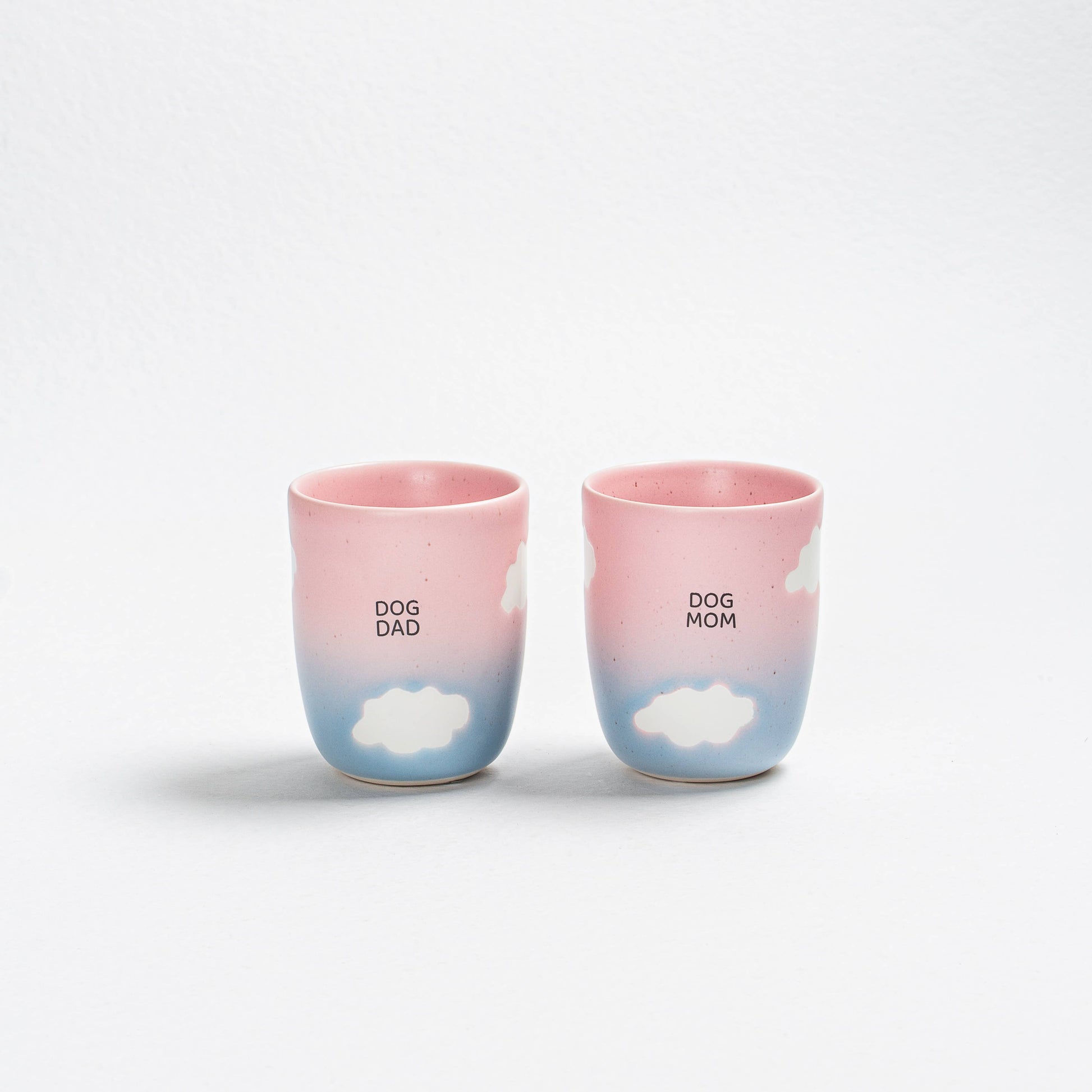 Two ceramic cups with 'DOG DAD' and 'DOG MOM' text and cloud designs on a white background.