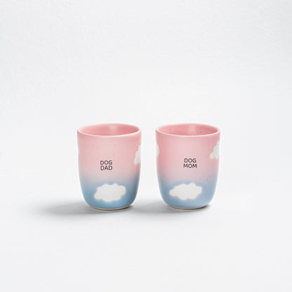 Two ceramic cups with 'DOG DAD' and 'DOG MOM' text and cloud designs on a white background.