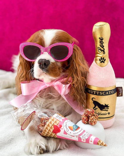 Dog wearing pink sunglasses with a bottle of 'Muttscato' and a toy ice cream cone on a pink background