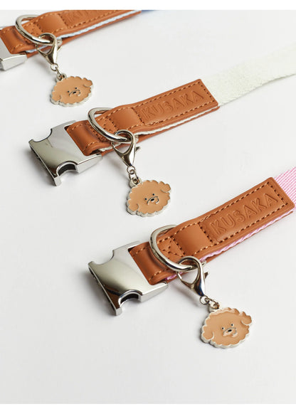 Three tan faux-leather strap accessories with silver metal buckles and hanging lion-face charms, arranged diagonally on a white background. The center strap is white, and the bottom-right strap is light pink.