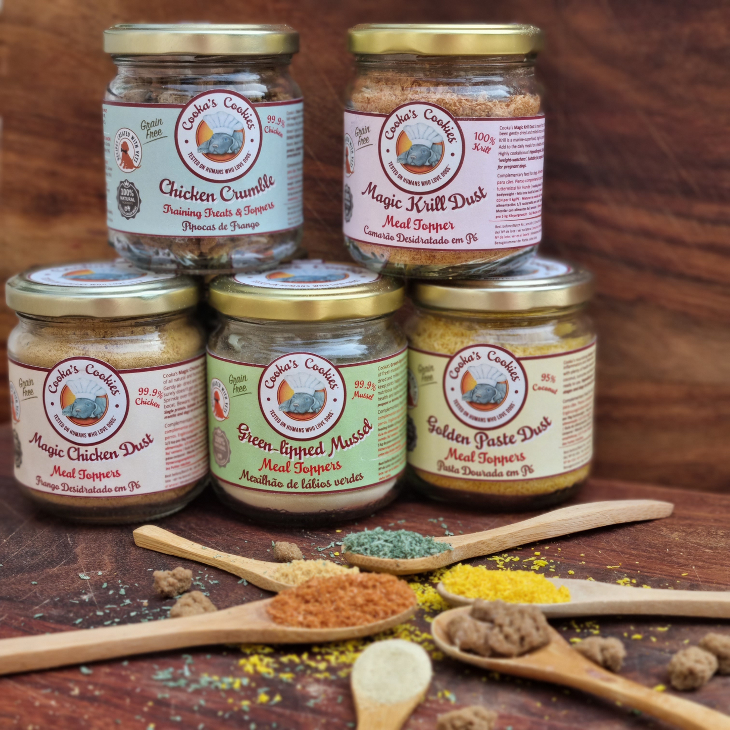 Jars of 'Cookin' Coosie' pet food products on a wooden surface with wooden spoons containing different ingredients.