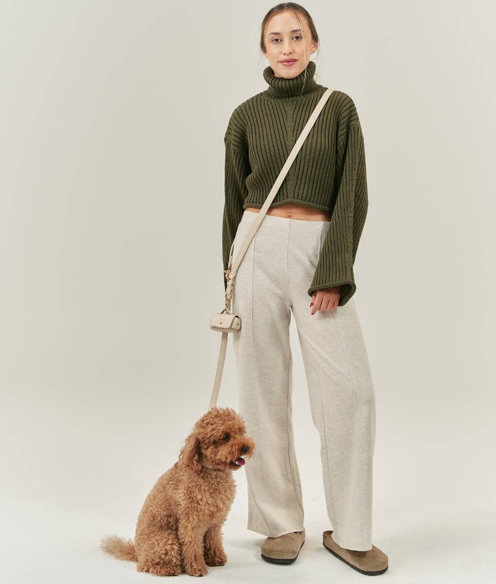 Woman in green sweater and beige pants standing next to a small brown dog on a white background