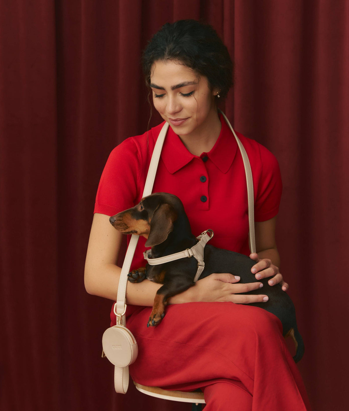 Woman in a red outfit holding a small dog with a leash against a red curtain background