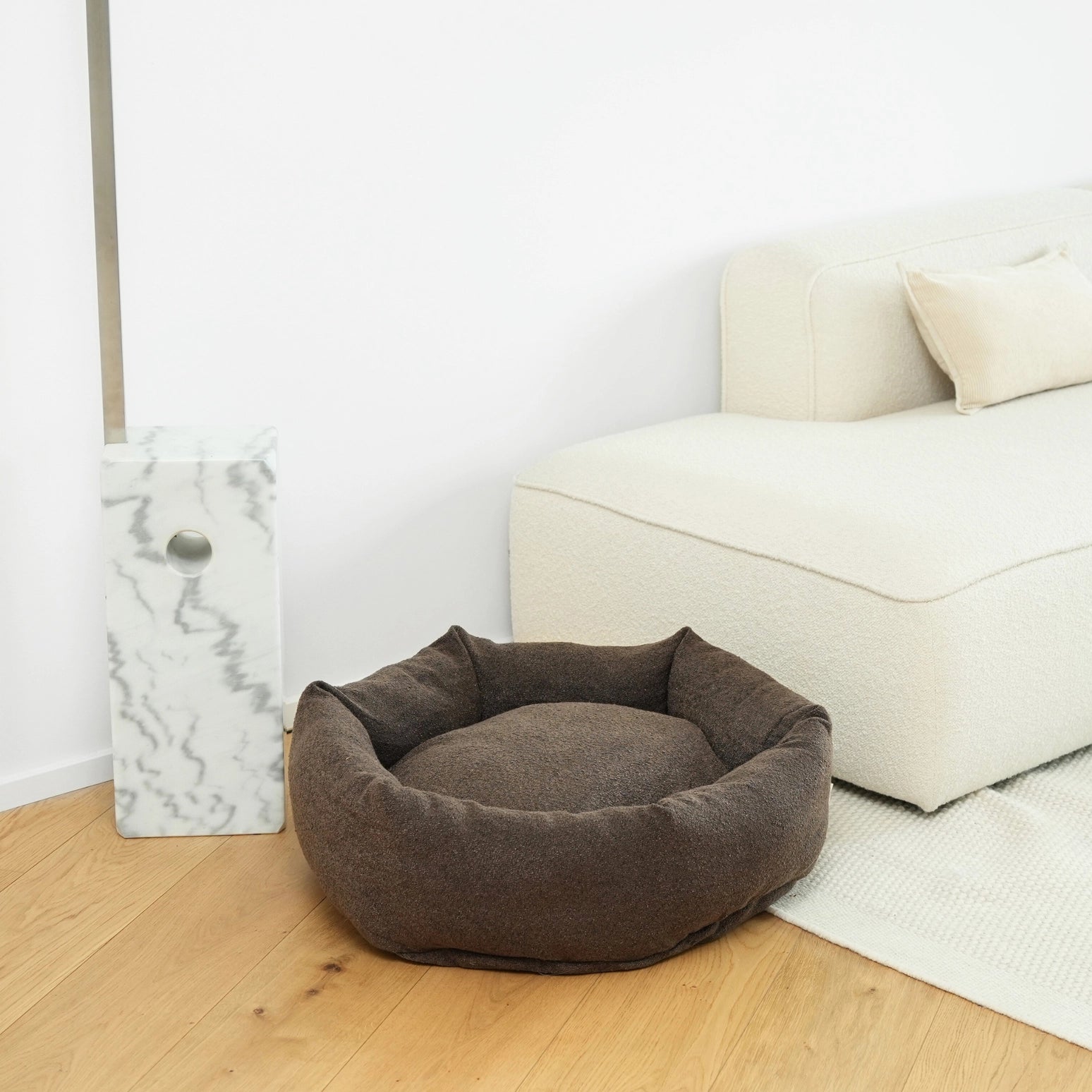 Brown pet bed on a wooden floor with a white wall and sofa in the background