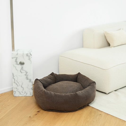 Brown pet bed on a wooden floor with a white wall and sofa in the background