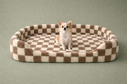 A small chihuahua sits centered in a plush oval pet bed with a brown and cream checkered pattern, set against a solid sage green background.