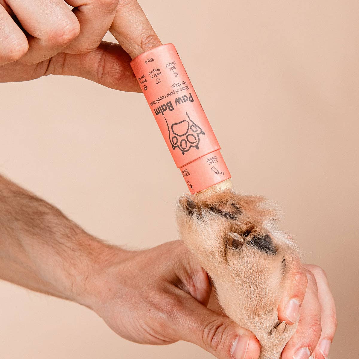 Person applying a pink paw balm to a dog's paw on a beige background