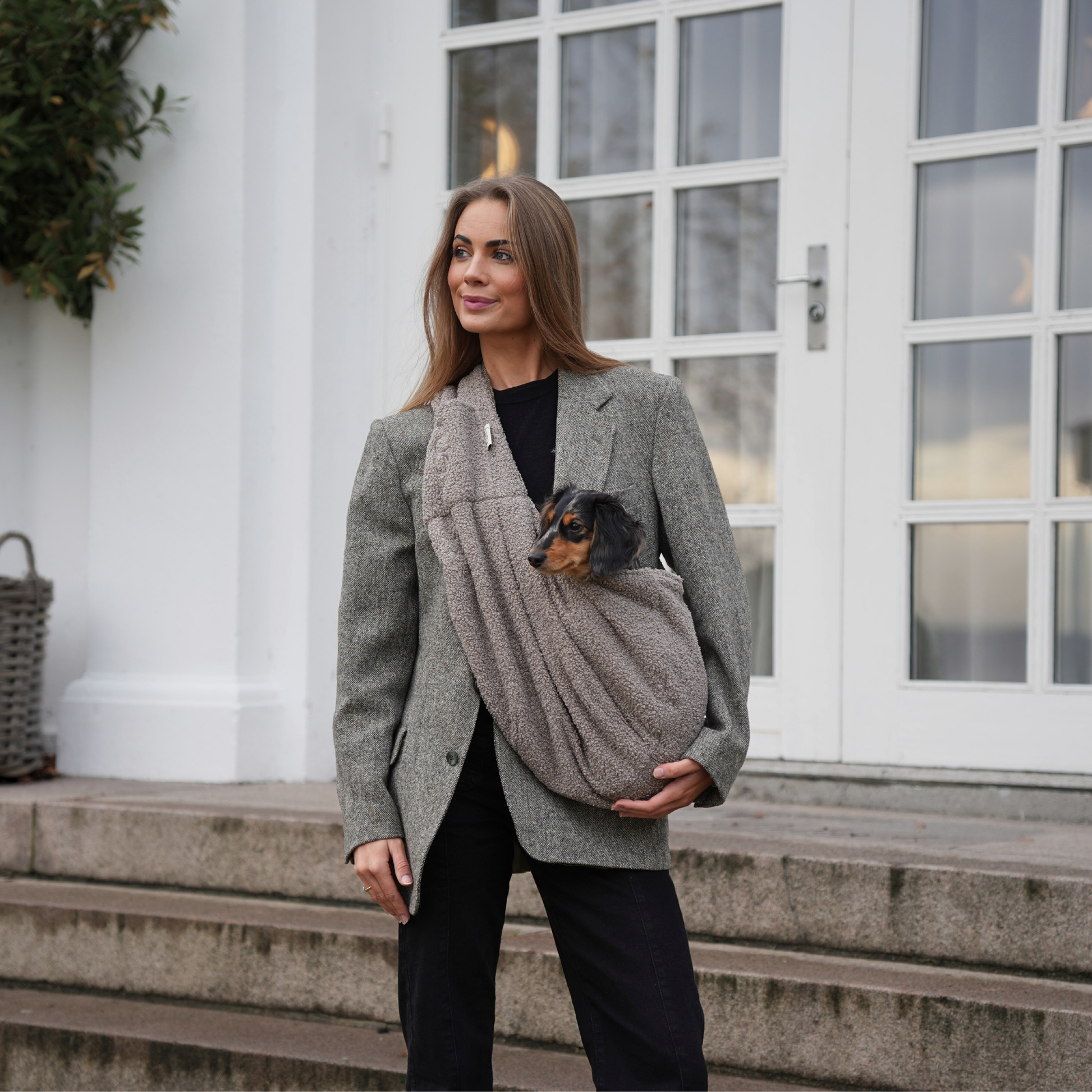 Woman holding a small dog in a sling while standing outdoors.