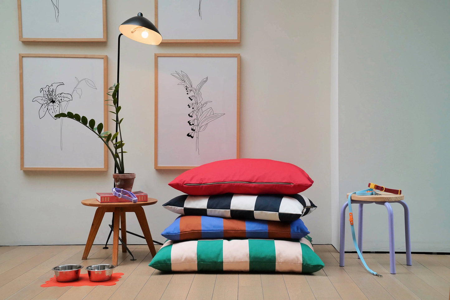 Stack of colorful cushions on a wooden floor with framed artwork and a lamp in the background.