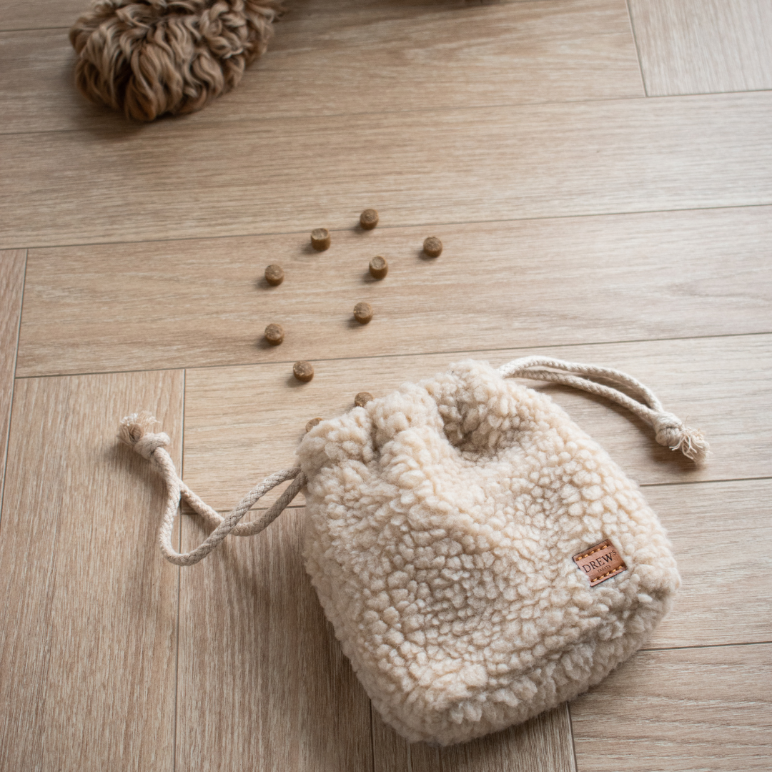 Beige sherpa drawstring bag on a wooden floor with scattered dog treats.