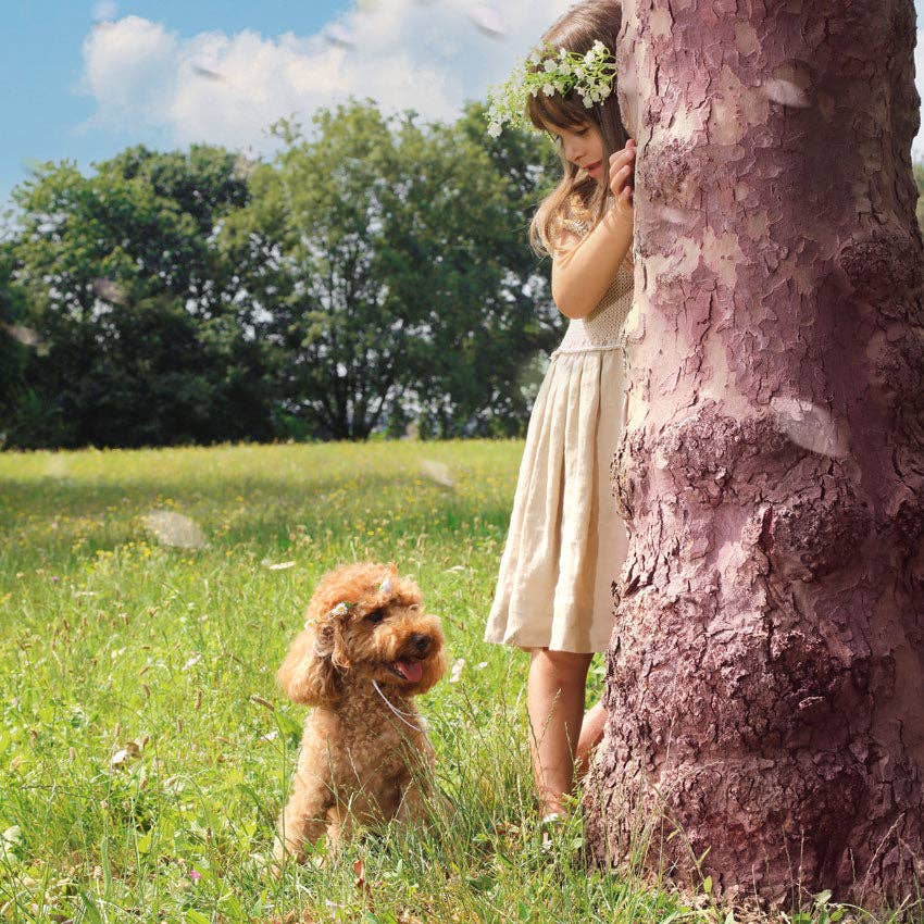 Girl in a dress standing next to a tree with a dog in a grassy field